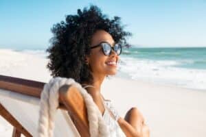 Happy young black woman relaxing on deck chair at beach wearing spectacles